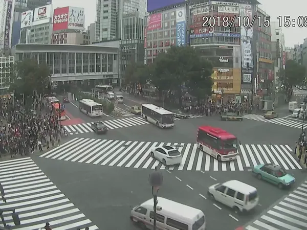 The Shibuya Scramble Crossing seen from above, with a red bus, a taxi and a white van crossing
