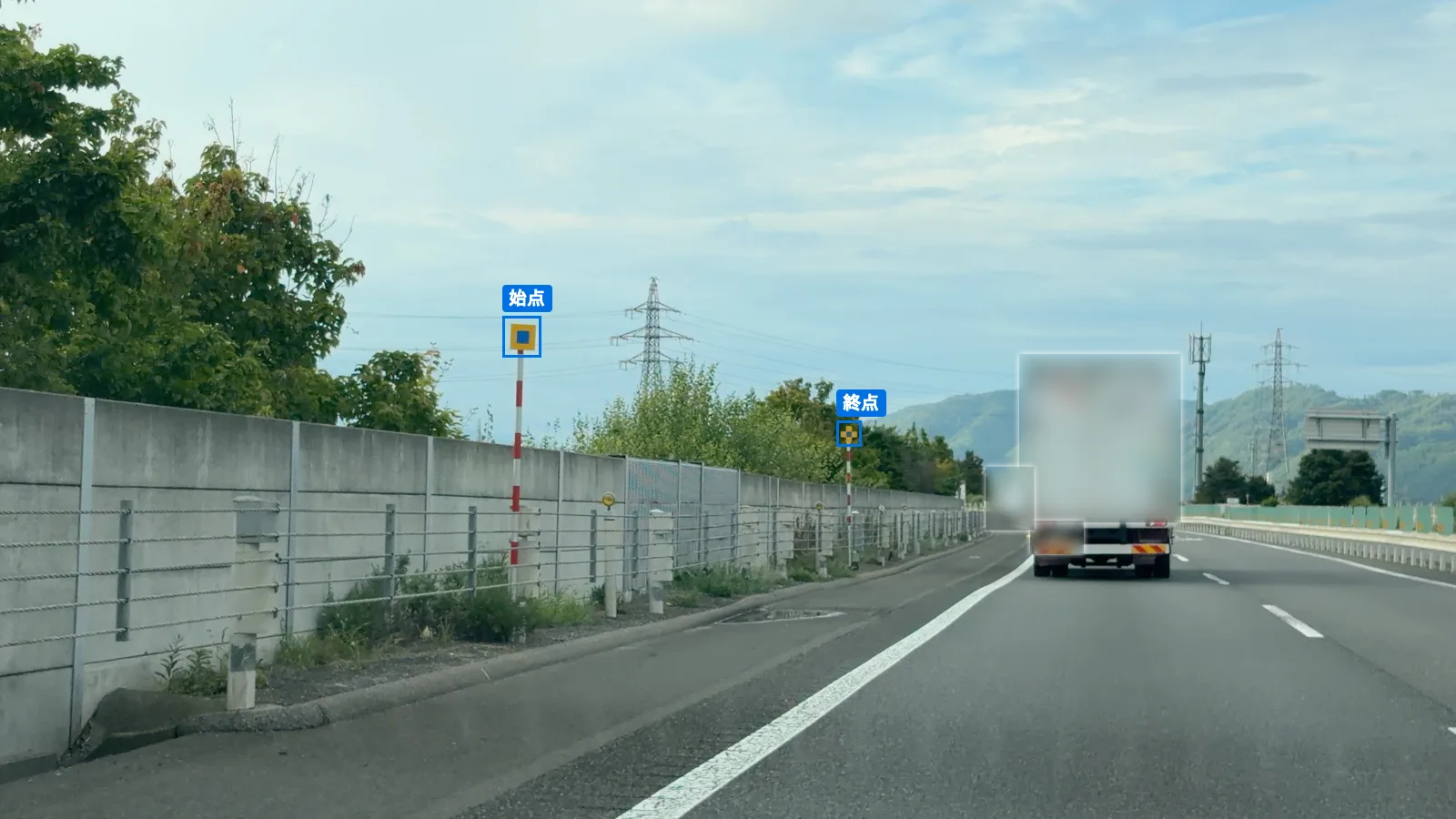 高速道路の左の路肩に立つ投雪防止表示板。手前の始点（黄色の正方形の中に青い正方形）と、遠くの終点（黄色と青の十字形）が青い枠で囲まれている。前を走るトラックはぼかしてある