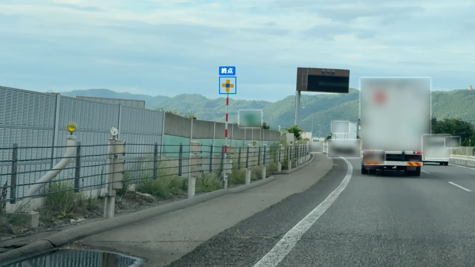 路肩の柵の奥に立つ、黄色と青の十字形の終点の表示板が青い枠で囲まれている。車両と案内標識はぼかしてある